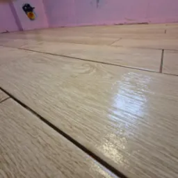 Świeżo ułożone, błyszczące, beżowe płytki podłogowe imitujące drewno w łazience, widoczne przyłącze wodne w ścianie z różową hydroizolacją.