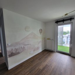 G-WORK Gaspar Gontarz - Pokój z brązowymi panelami podłogowymi i ścianą ozdobioną malowidłem przedstawiającym pejzaż z górą, słońcem i balonem, grzejnik pod oknem balkonowym.