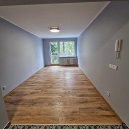 G-WORK Gaspar Gontarz - Pusty pokój po remoncie z drewnianą podłogą, szarymi ścianami i białymi listwami, widok na okno z grzejnikiem i balkonem.