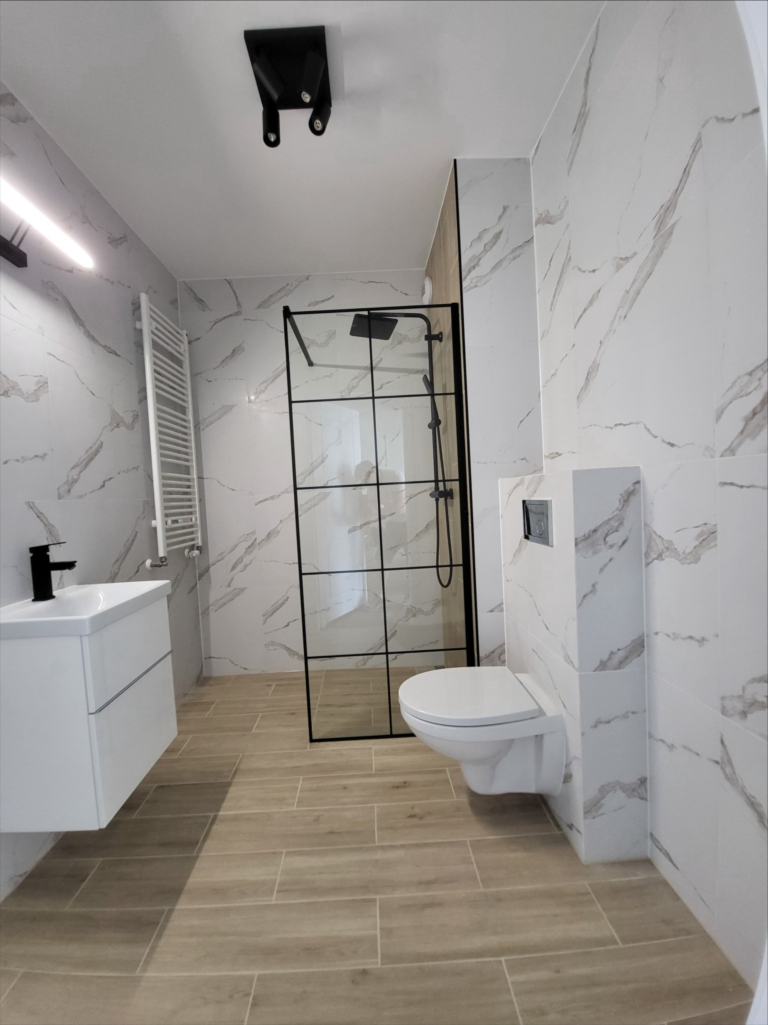 Nowoczesna łazienka z kabiną prysznicową w czarnej ramie, wiszącą toaletą i umywalką z czarną armaturą, wykończona płytkami imitującymi marmur i drewno, oświetlona punktowym światłem.