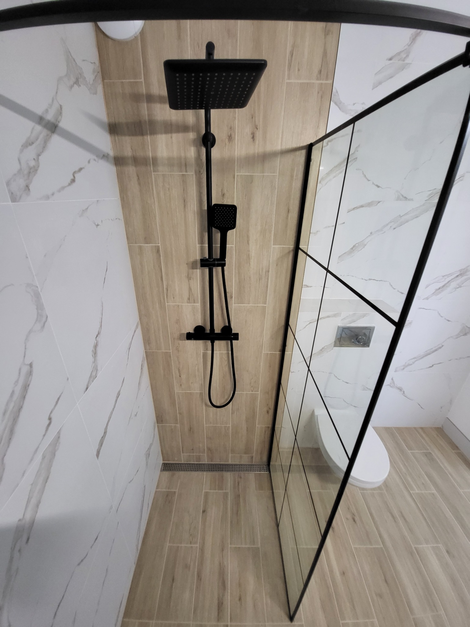 Nowoczesna kabina prysznicowa typu walk-in z czarną armaturą, wykończona płytkami imitującymi drewno i marmur, z widocznym odpływem liniowym i toaletą w tle.