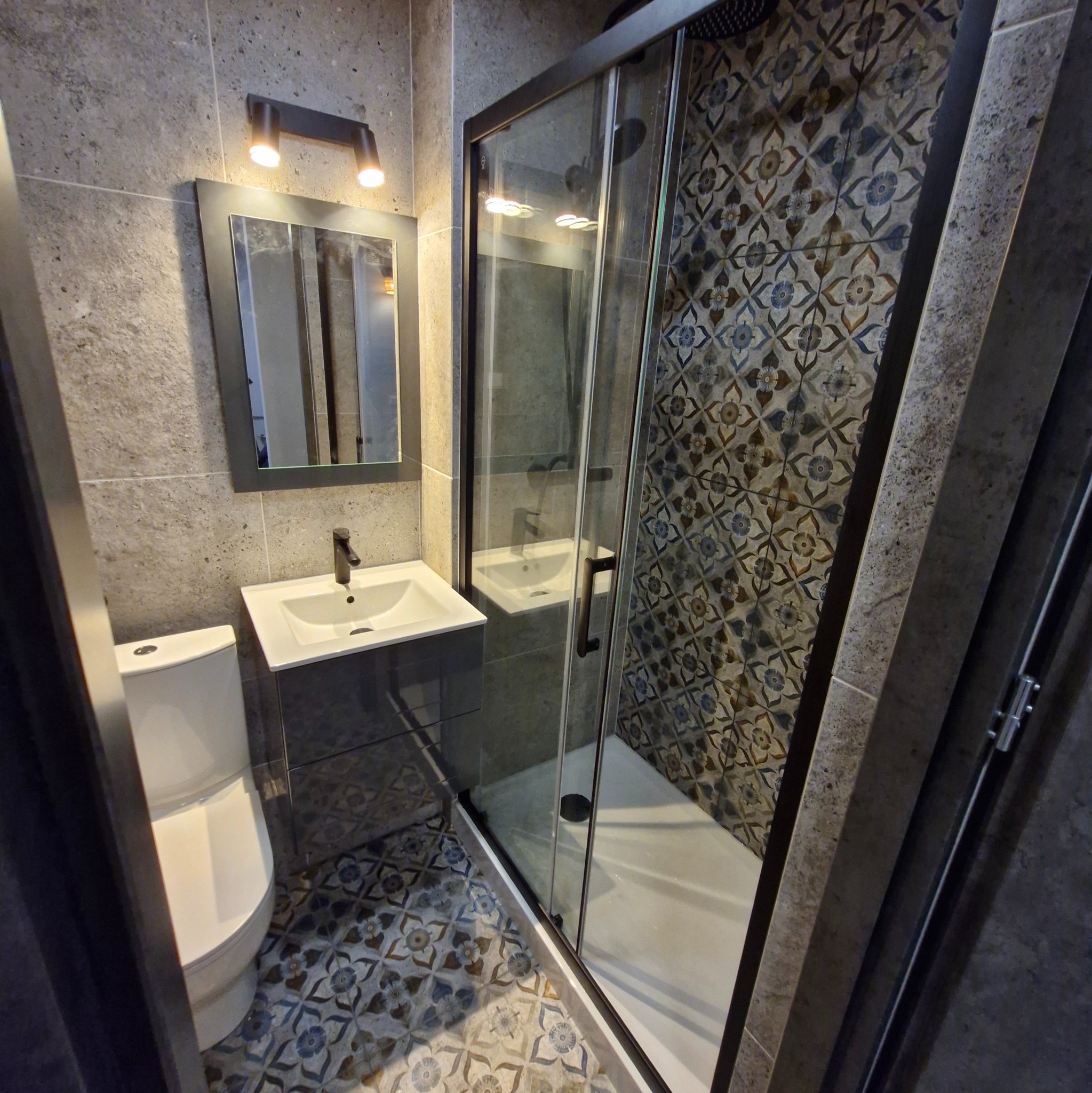 Nowoczesna łazienka z kabiną prysznicową z czarną ramą, umywalką z szafką w kolorze grafitowym, toaletą i lustrem podświetlanym lampą punktową, wykończona płytkami w odcieniach szarości...