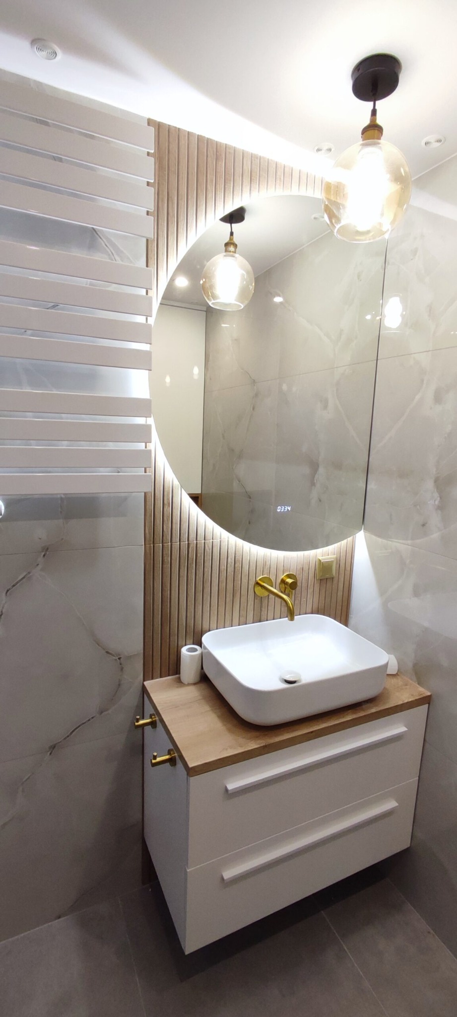 Nowoczesna łazienka z okrągłym lustrem podświetlanym LED, umywalką nablatową na drewnianym blacie i złotą armaturą, marmurowe płytki na ścianach, biały grzejnik drabinkowy.