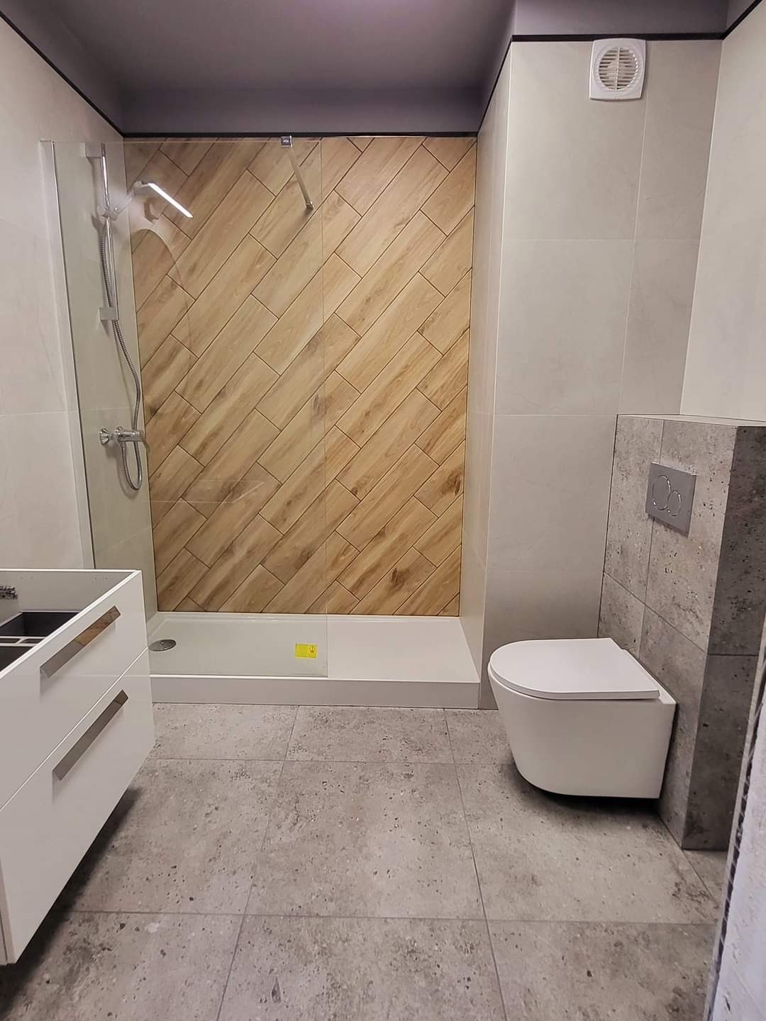 Nowoczesna łazienka z drewnopodobnymi płytkami ułożonymi w jodełkę pod prysznicem, szare płytki na ścianach, biała wisząca toaleta i niedokończona szafka pod umywalkę.