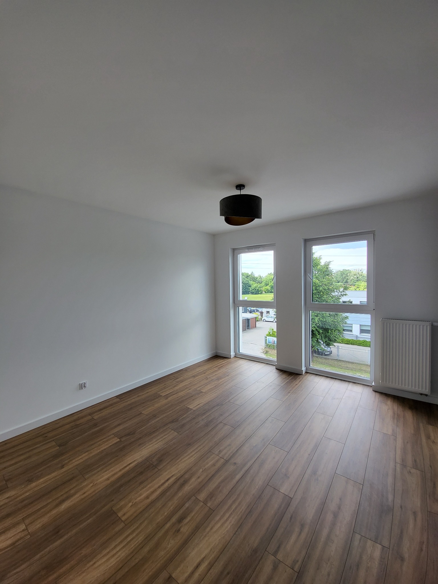 Puste, jasne pomieszczenie z drewnopodobną podłogą, białymi ścianami i dwoma oknami z widokiem na zieleń, uzupełnione czarną lampą sufitową i grzejnikiem.