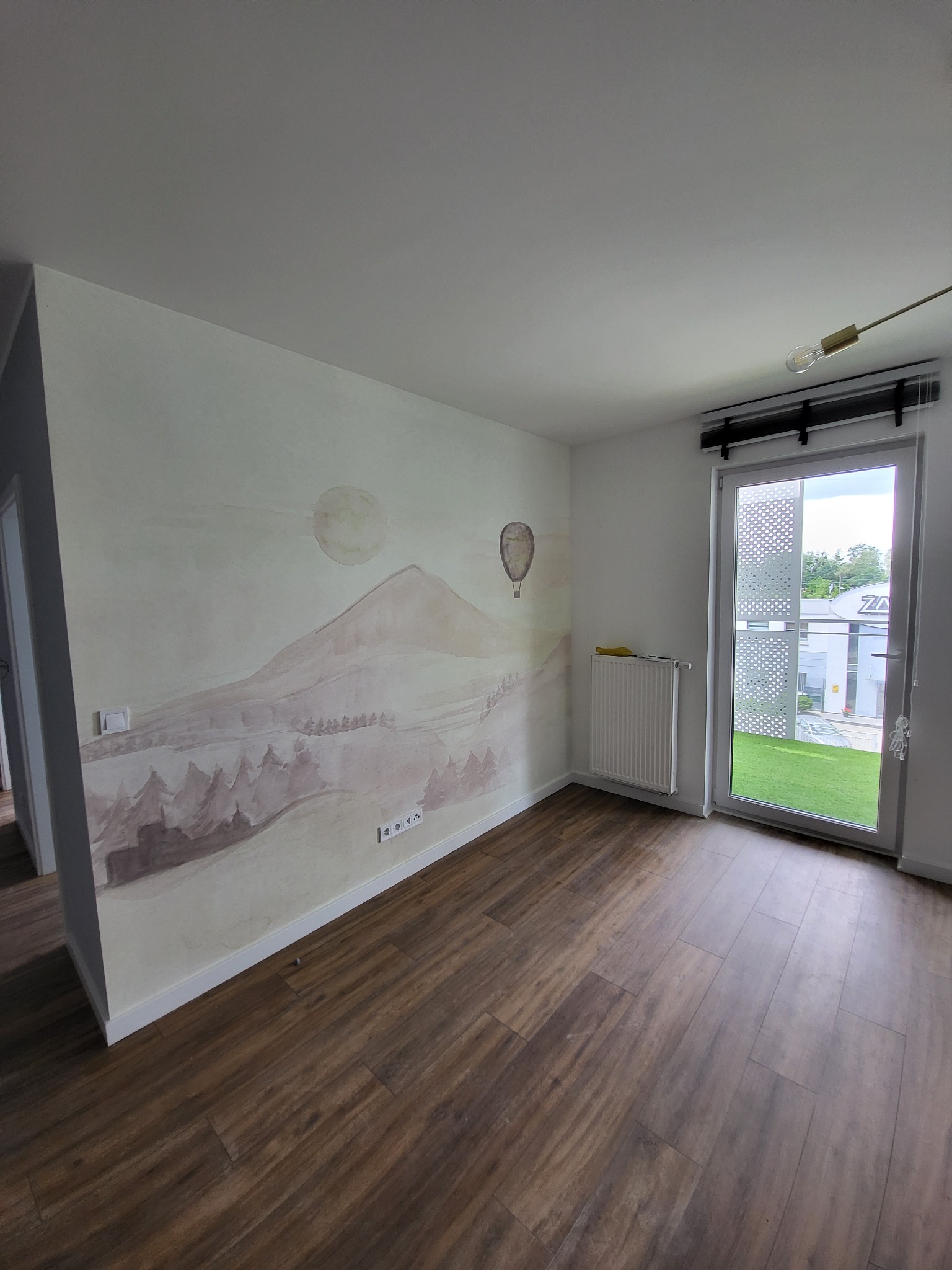 Pokój z brązowymi panelami podłogowymi i ścianą ozdobioną malowidłem przedstawiającym pejzaż z górą, słońcem i balonem, grzejnik pod oknem balkonowym.