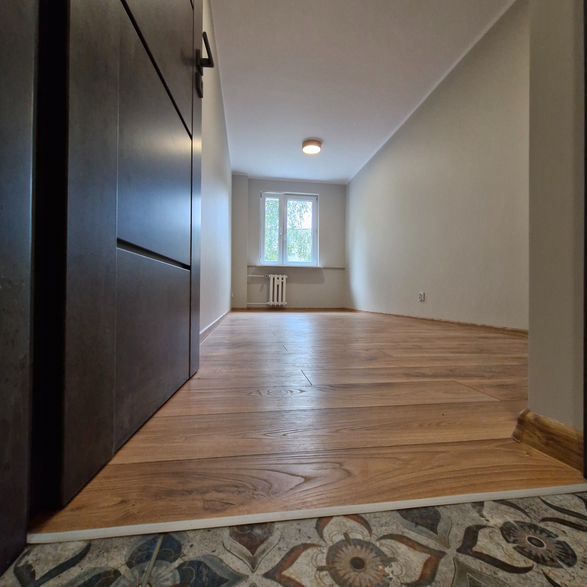 Ukończone układanie paneli podłogowych z drewnopodobnym wzorem, widoczne przejście z płytek o wzorze kwiatowym, fragment ciemnej szafy w tle, pokój z oknem i grzejnikiem.