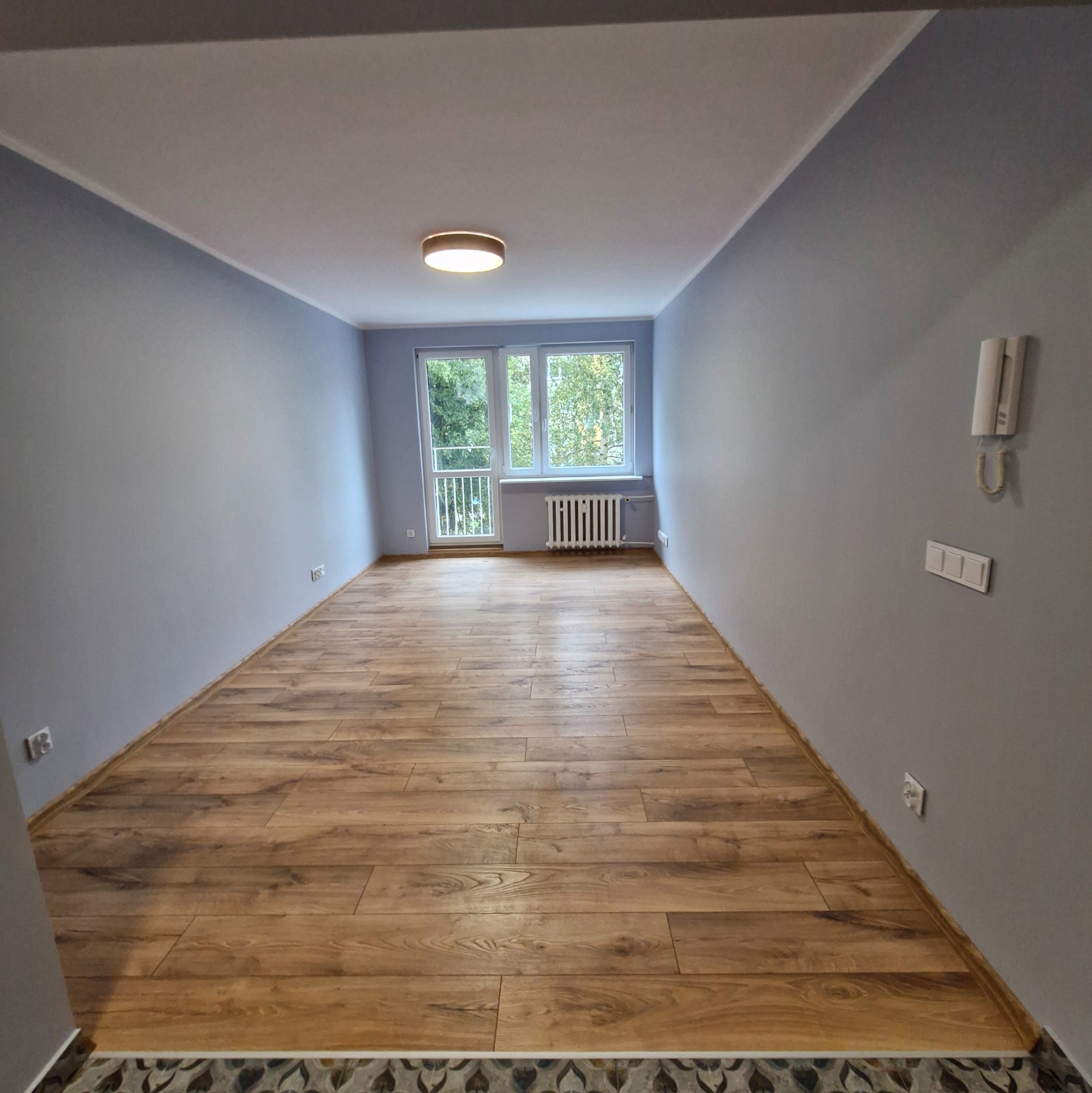 Pusty pokój po remoncie z drewnianą podłogą, szarymi ścianami i białymi listwami, widok na okno z grzejnikiem i balkonem.