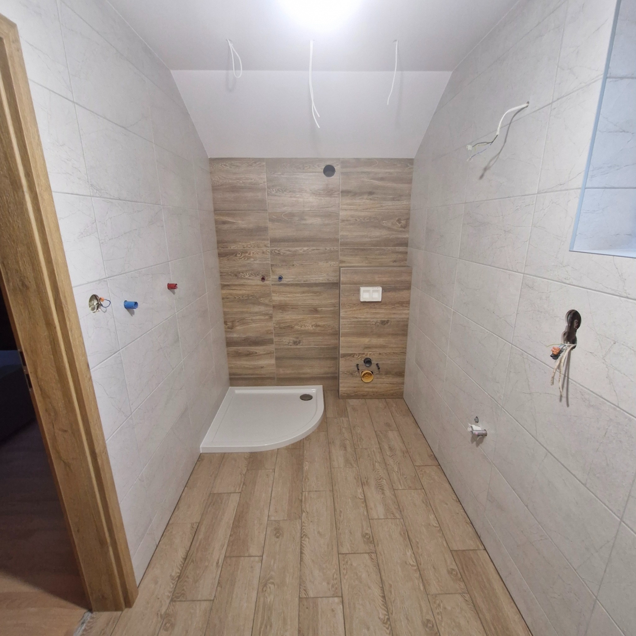 Wnętrze łazienki w trakcie remontu: ułożone płytki imitujące drewno na podłodze i ścianach, biały brodzik, instalacja sanitarna i elektryczna w ścianach.