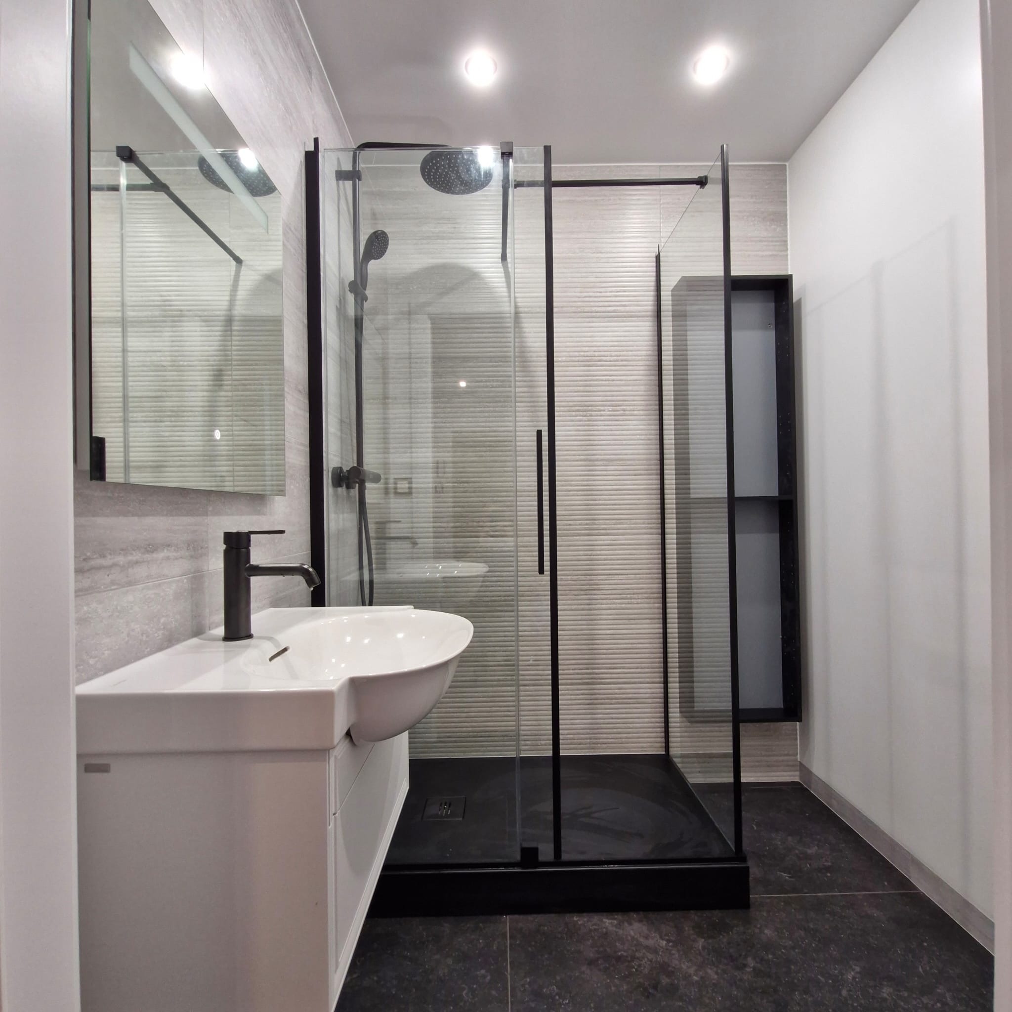 Nowoczesna łazienka z czarną armaturą i kabiną prysznicową typu walk-in, wykończona płytkami w odcieniach szarości z subtelną fakturą.