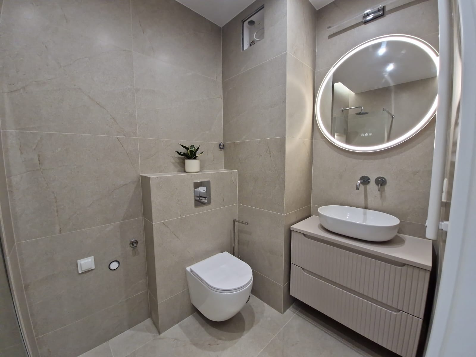 Nowoczesna łazienka w beżowych tonacjach. Uwagę przykuwa okrągłe lustro z podświetleniem LED nad umywalką nablatową i wisząca toaleta z rośliną.