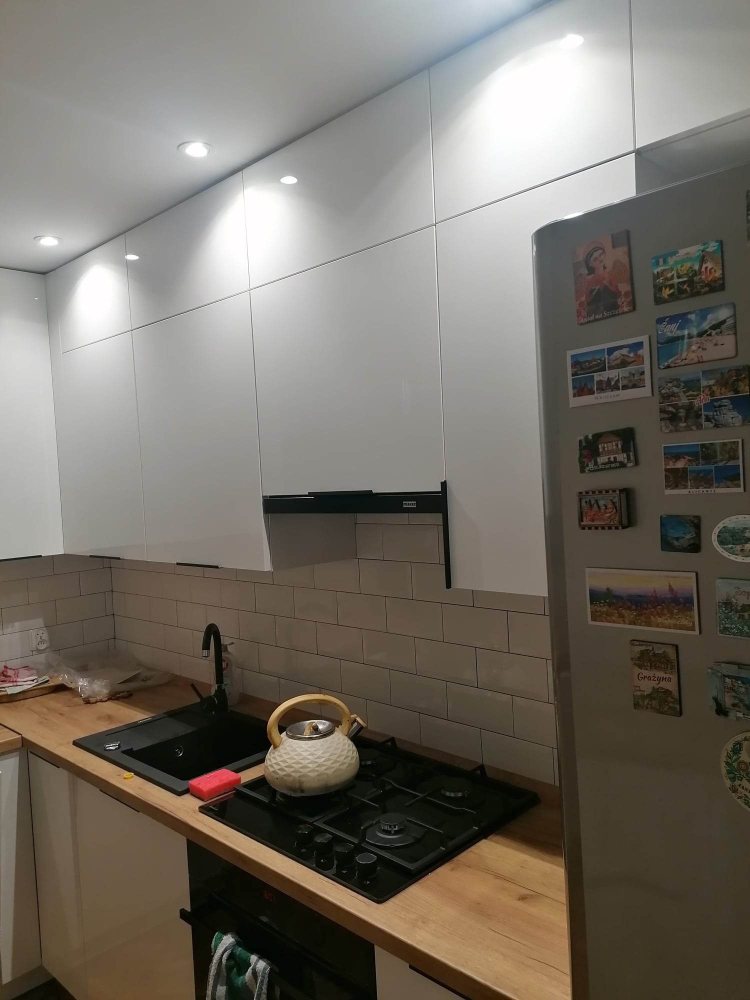 Białe szafki kuchenne z drewnianym blatem, czarnym zlewem i płytą gazową, na której stoi czajnik. Na lodówce widoczne magnesy z podróży.