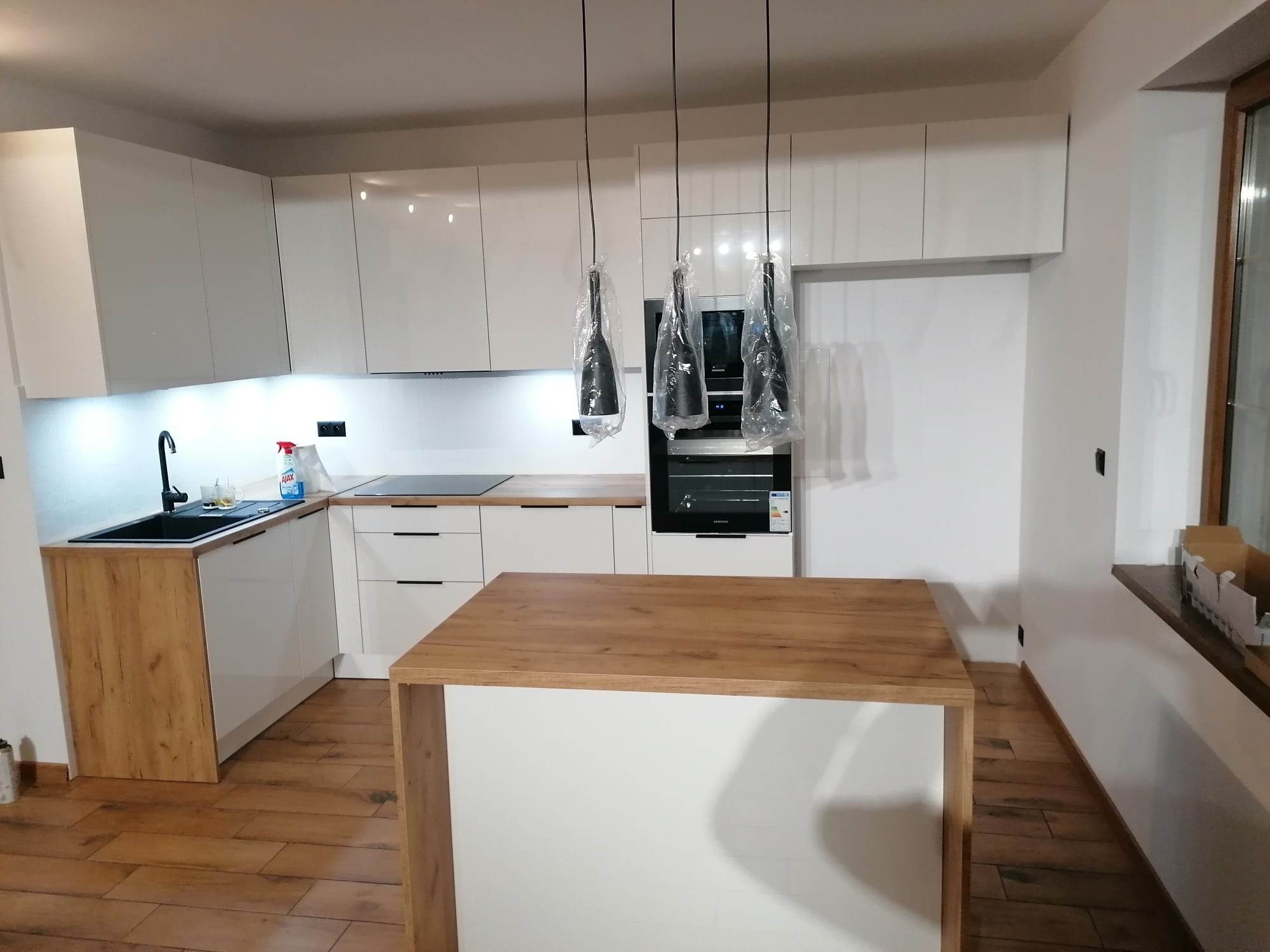 Nowoczesna, jasna kuchnia z wyspą w Olsztynie, z białymi, błyszczącymi frontami szafek i drewnianymi akcentami, lampy wiszące jeszcze w folii ochronnej, drewniana podłoga.