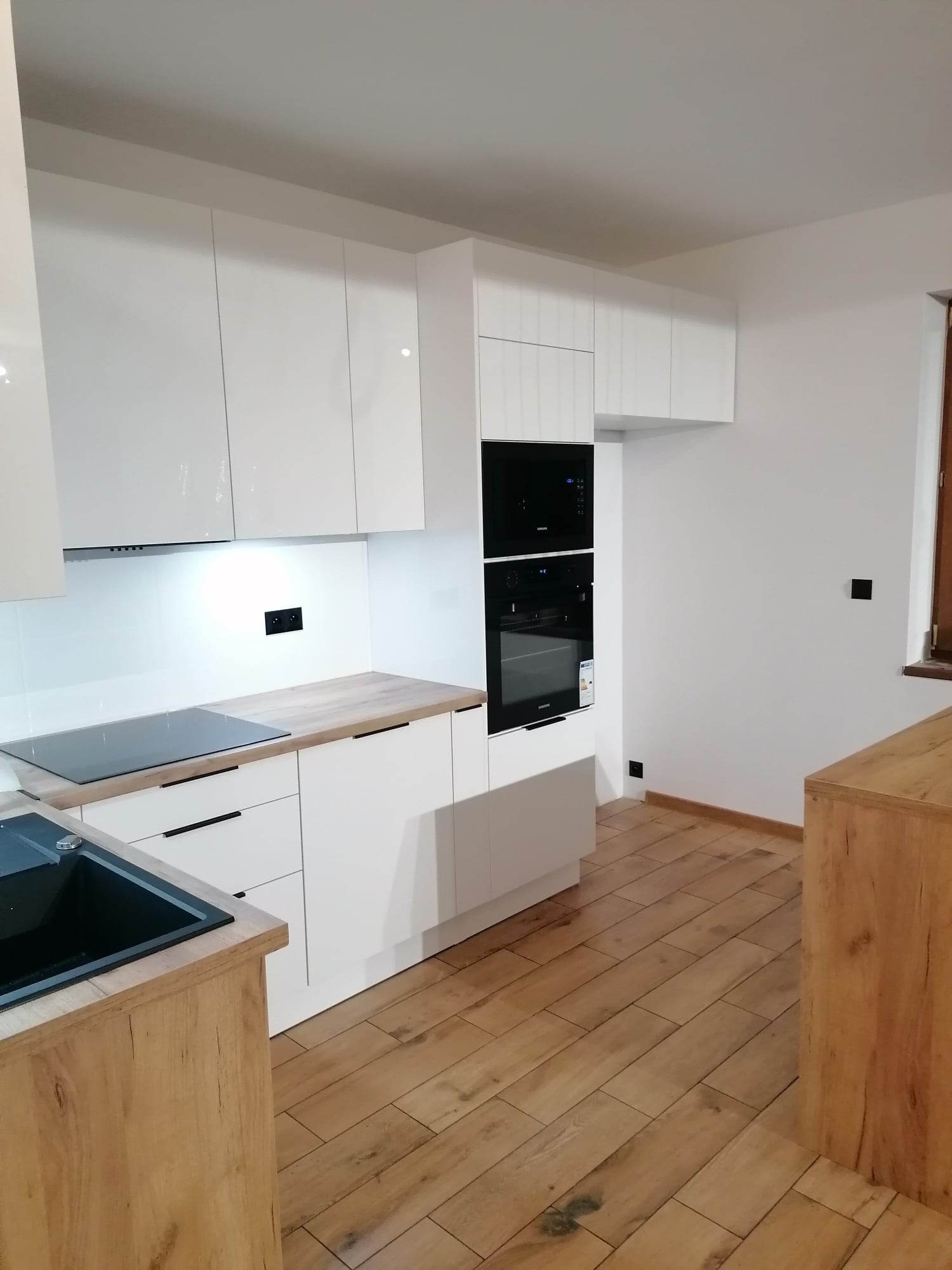 Nowoczesna kuchnia z białymi, lakierowanymi frontami, drewnianym blatem i podłogą w odcieniach brązu, z wbudowanym piekarnikiem i płytą indukcyjną.