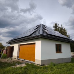 D&S Damian Ślęzak - Biały budynek gospodarczy z zamontowanymi panelami słonecznymi na dachu, pracownik wchodzący po drabinie, obok zaparkowany biały bus.