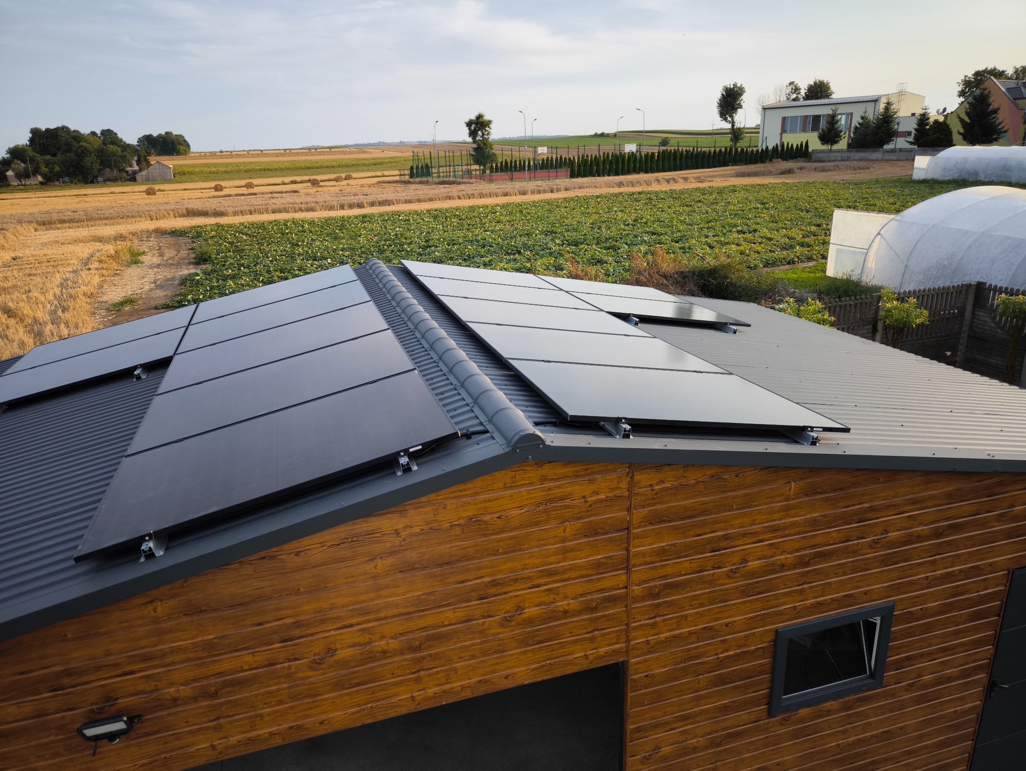 Panele słoneczne zamontowane na dachu budynku gospodarczego z drewnianą elewacją, w tle pole uprawne i zabudowania.