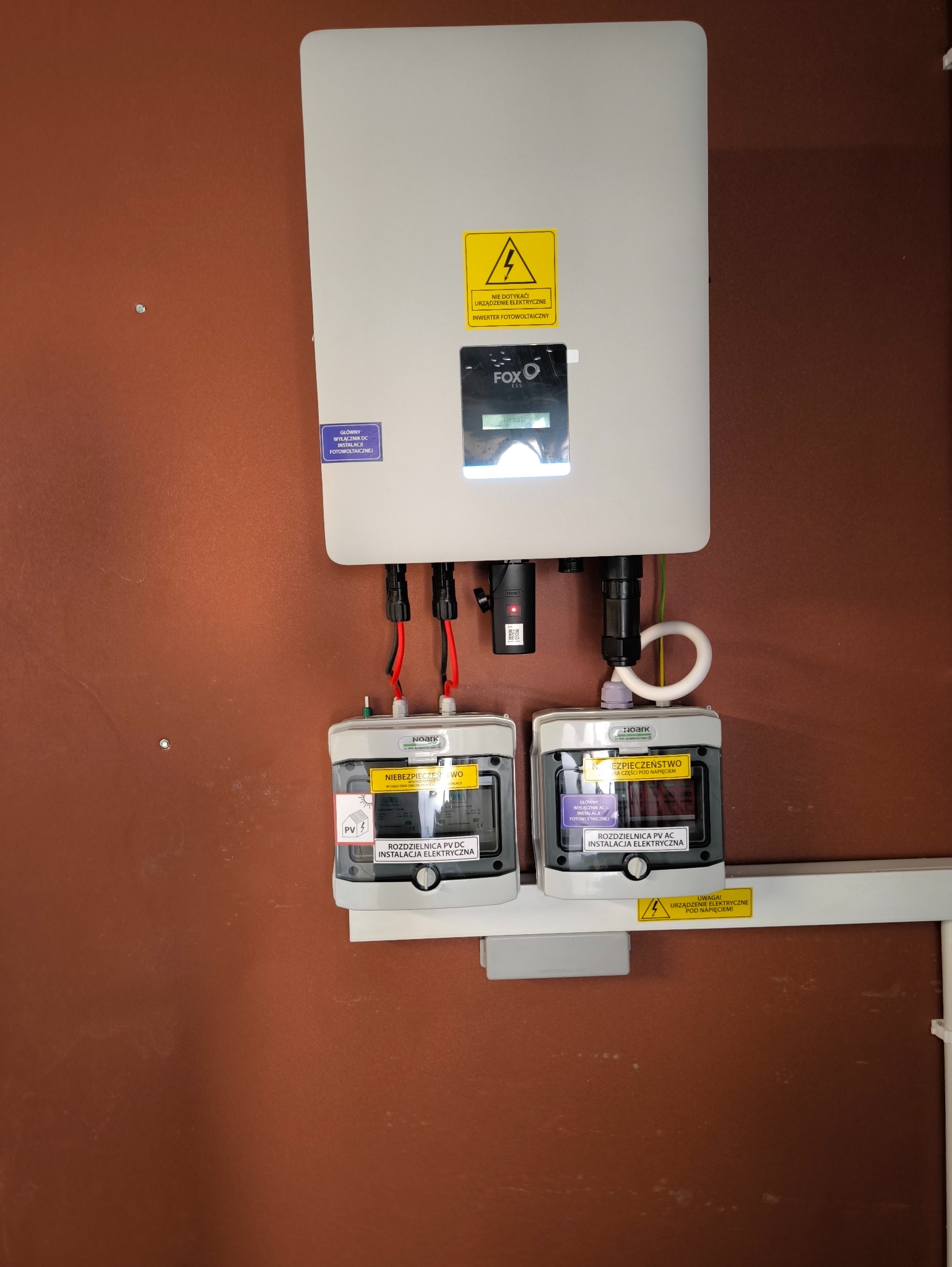 Zamontowany inwerter FOX ESS z rozdzielnicami PV DC i AC na ścianie o brązowym odcieniu, widoczne przewody i naklejki ostrzegawcze.