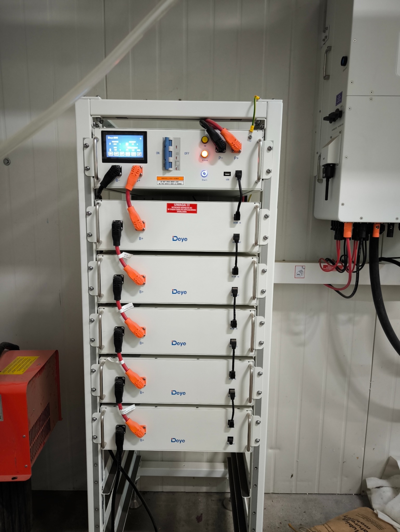 Regał z modułowymi akumulatorami Deye z podłączonymi kablami, obok widoczny inwerter, całość w pomieszczeniu o ścianach z blachy trapezowej.