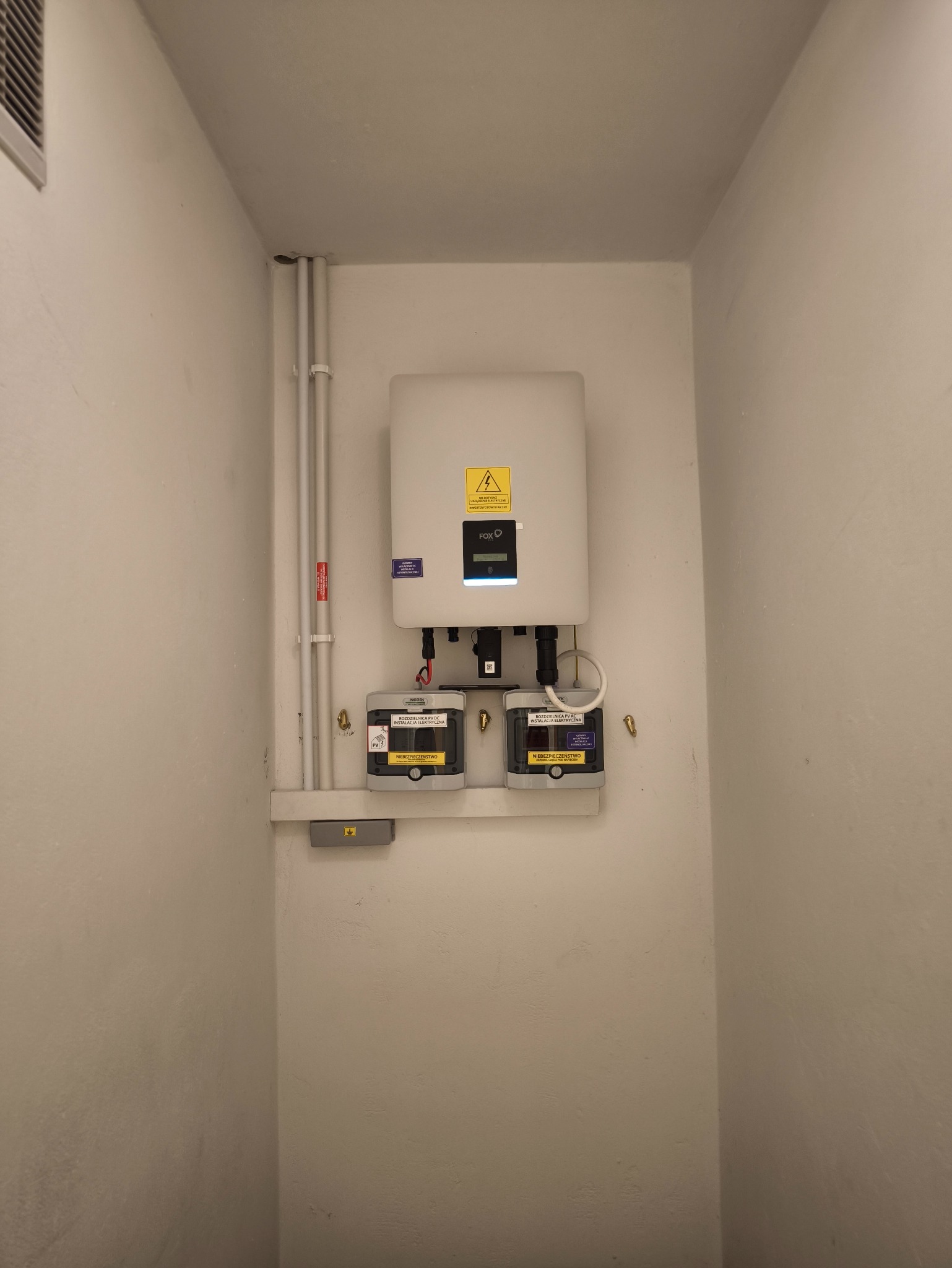 Biały inwerter fotowoltaiczny FOX z podłączonymi dwoma rozdzielnicami PV AC, zamontowany w rogu pomieszczenia z rurami instalacyjnymi na ścianie.