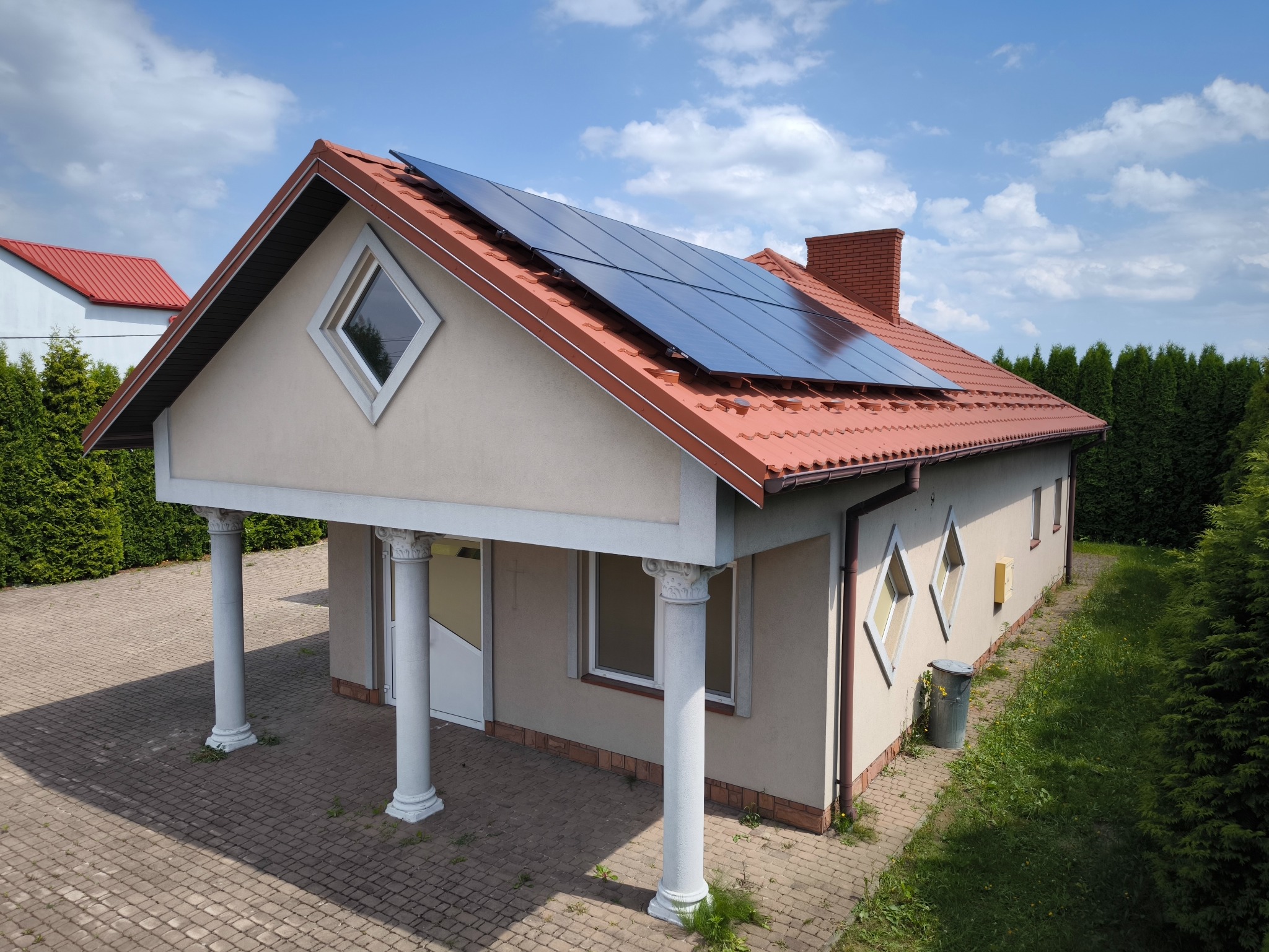 Dom jednorodzinny z czerwoną dachówką i panelami fotowoltaicznymi na dachu, widok z góry, błękitne niebo z chmurami.