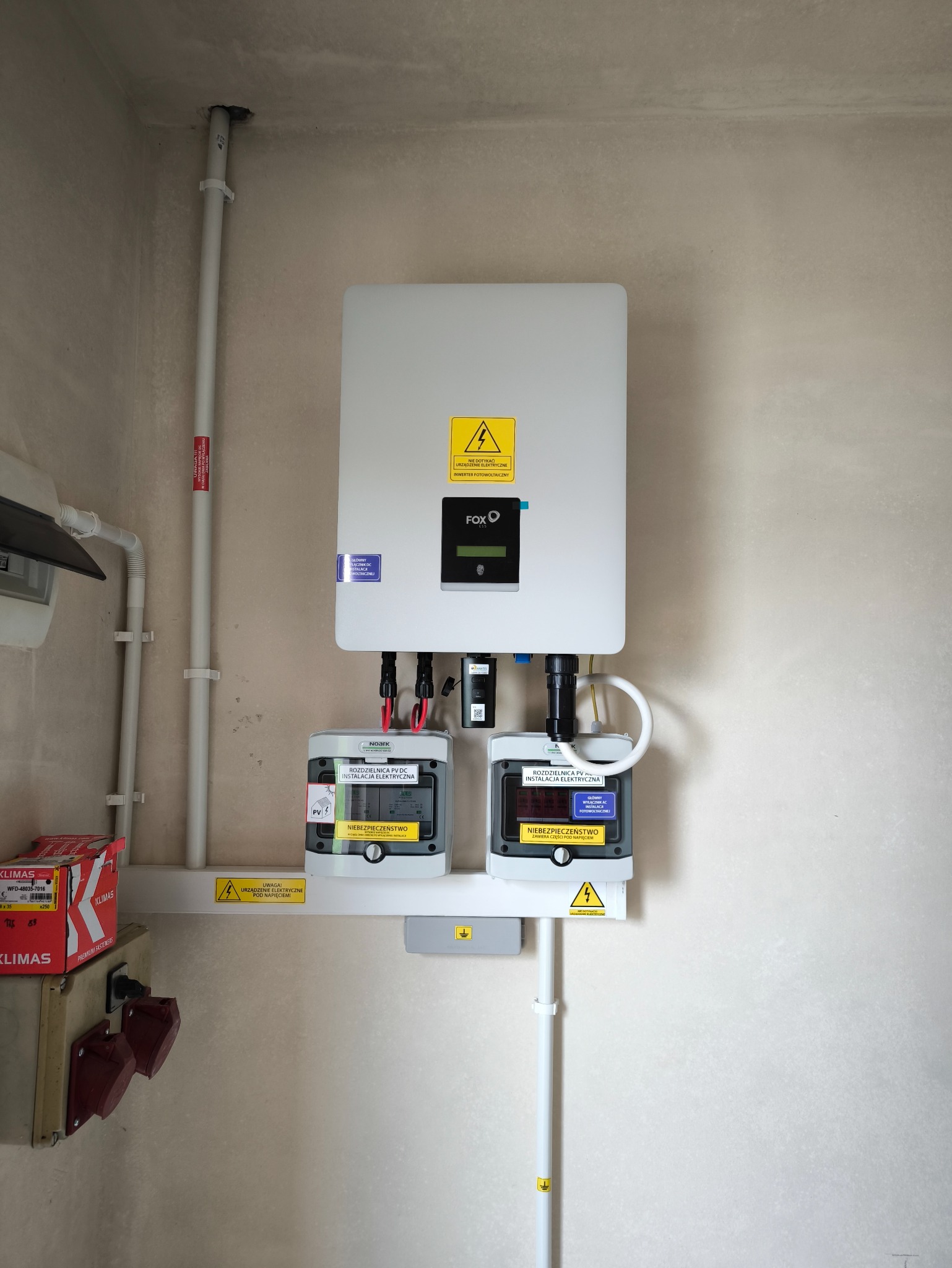 Zamontowany inwerter fotowoltaiczny Fox ESS z dwoma licznikami energii Noark PV DC na ścianie, widoczne przewody i oznaczenia ostrzegawcze.