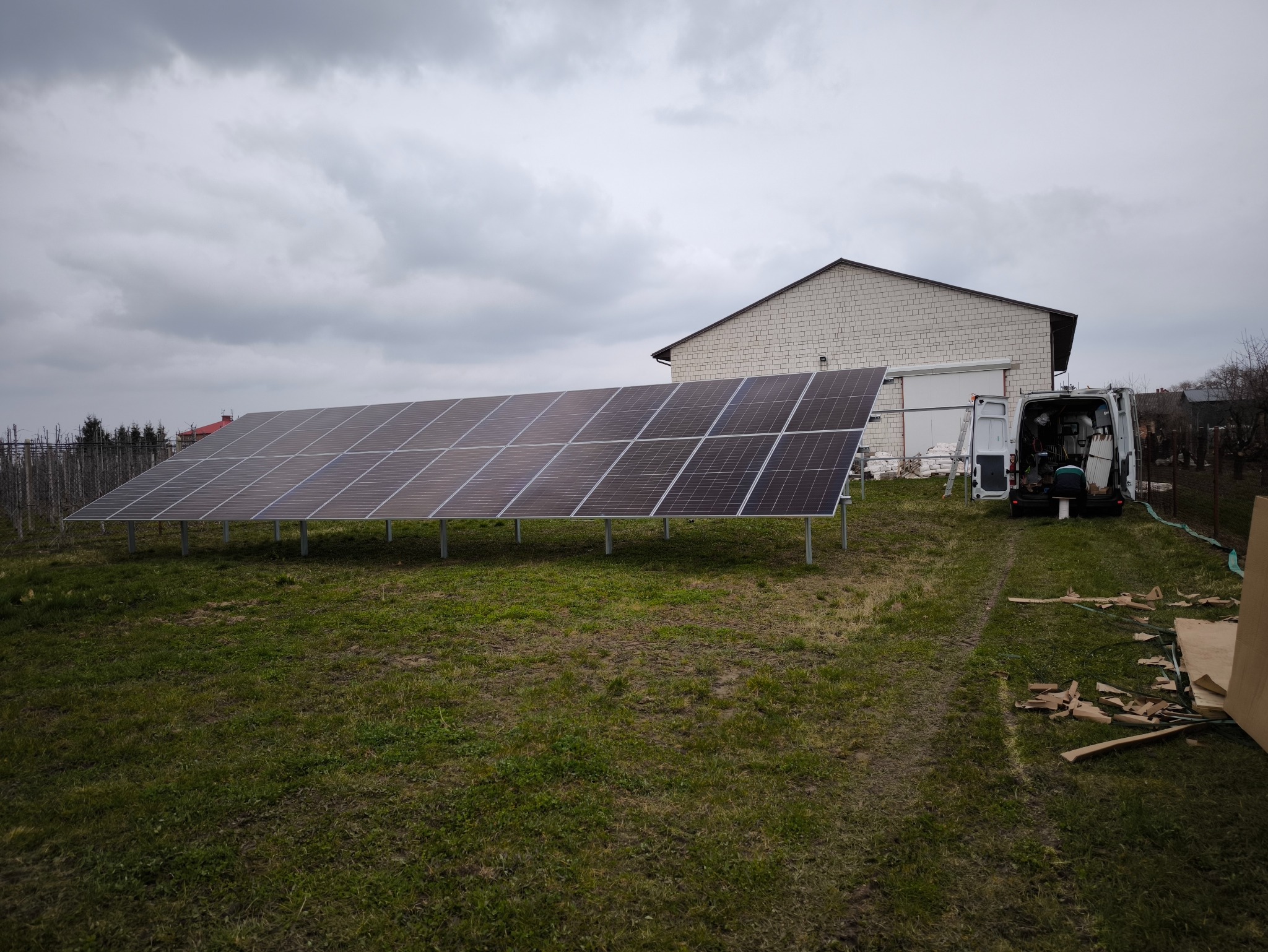Rząd paneli słonecznych na metalowych wspornikach, obok biały budynek gospodarczy i samochód dostawczy z otwartymi drzwiami, pochmurne niebo.