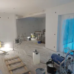 Wnętrze w trakcie remontu: ściany przygotowane do malowania, widoczne ślady szpachlowania, podłoga zabezpieczona folią, sprzęt do malowania natryskowego, drabina, worki z materiałami budowlanymi...