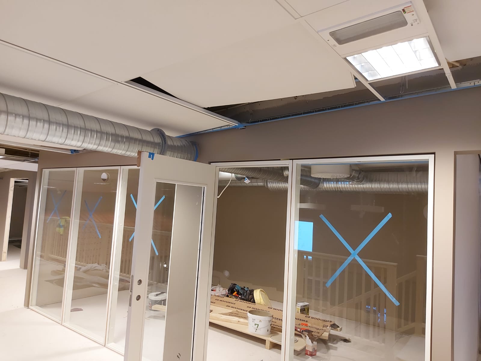 Wnętrze biurowe w trakcie prac wykończeniowych: widoczne przeszklone ścianki działowe zabezpieczone niebieską taśmą, odsłonięte rury wentylacyjne pod sufitem i rozłożone materiały budowlane.
