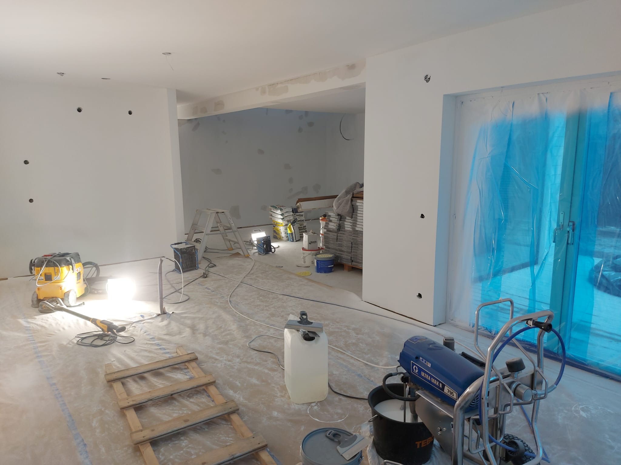 Wnętrze w trakcie prac przygotowawczych do malowania: zabezpieczona folią stolarka okienna, zakryta podłoga, ściany z widocznymi punktami po gniazdkach elektrycznych, szpachlowane ubytki w ścianach...