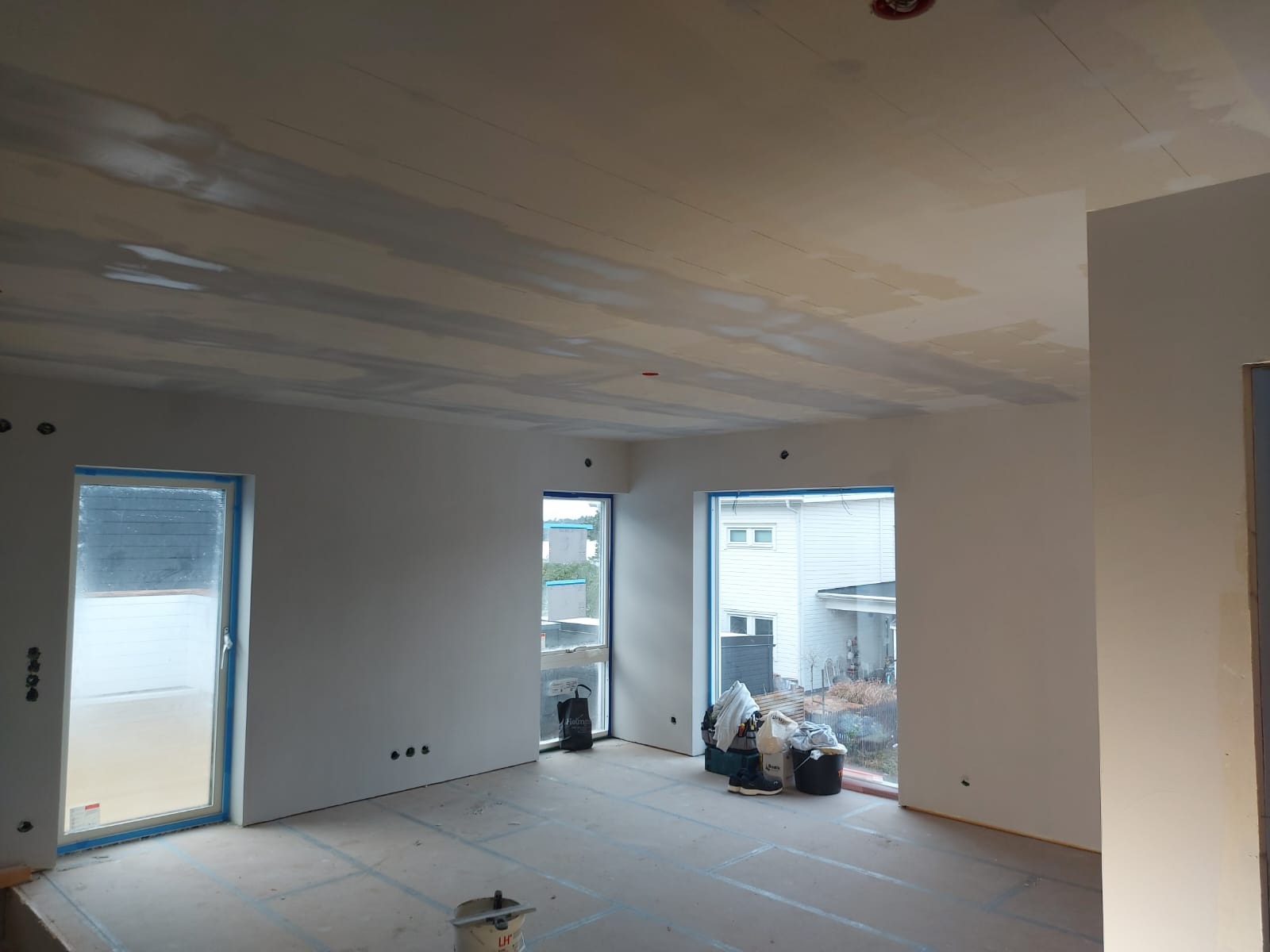 Pokój w trakcie szpachlowania sufitu z widocznymi śladami szpachli, zabezpieczone okna taśmą malarską, przy oknach worki z materiałami budowlanymi, na podłodze folia ochronna