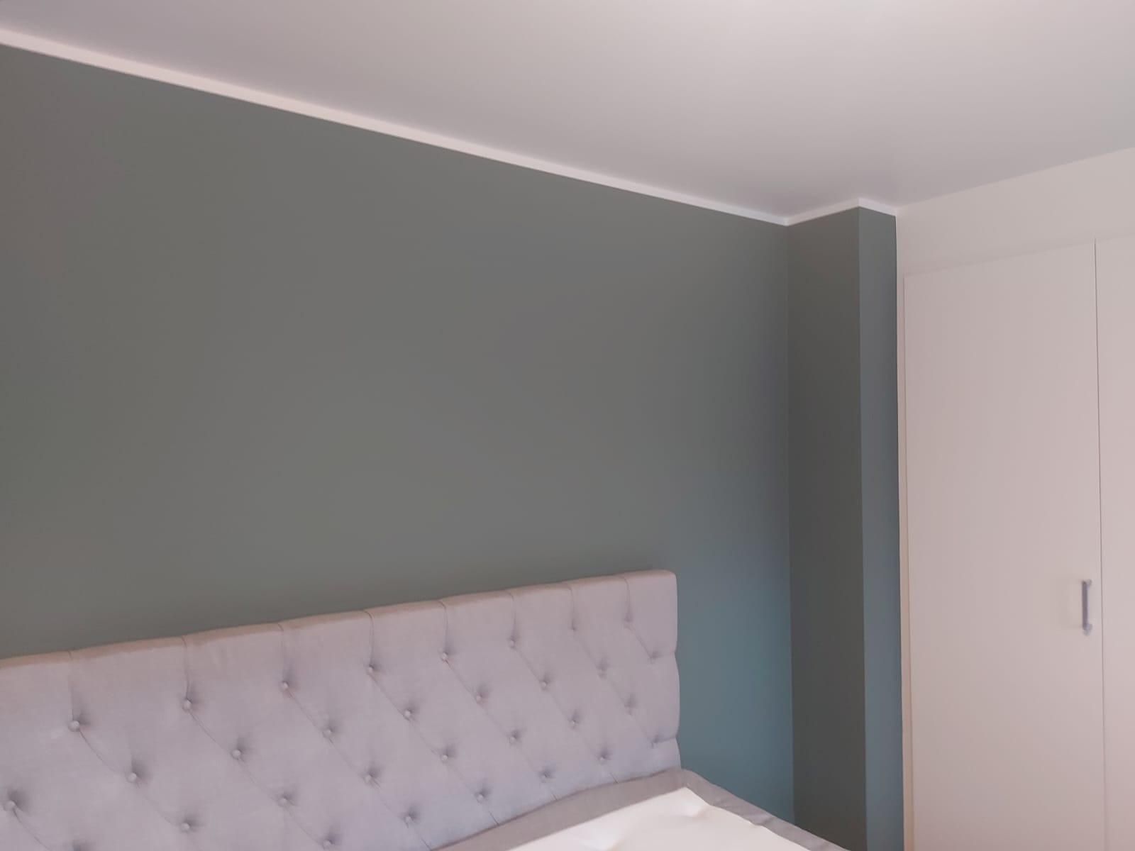 Pomieszczenie po odświeżeniu: ściana w odcieniach szarości, widoczna wezgłowie łóżka z pikowaniem i biała szafa w tle.