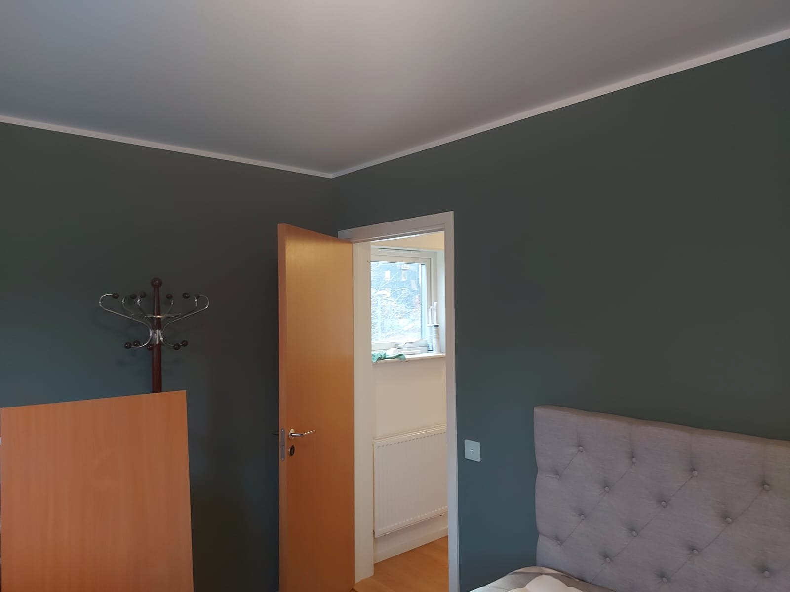 Ściana w kolorze ciemnej zieleni, świeżo pomalowana, z widocznym fragmentem łóżka z wezgłowiem, otwarte drzwi do jasnego pomieszczenia z oknem, w rogu stoi wieszak na ubrania.