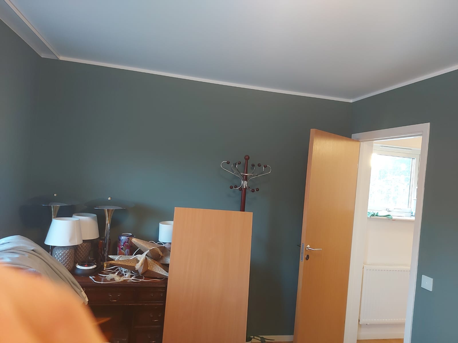 Pokój przed remontem: ciemnozielona ściana, niebieskawo-biały sufit z listwą, drewniane drzwi uchylone, widoczny fragment okna, biurko z lampami i dekoracjami, w tle wieszak na ubrania.