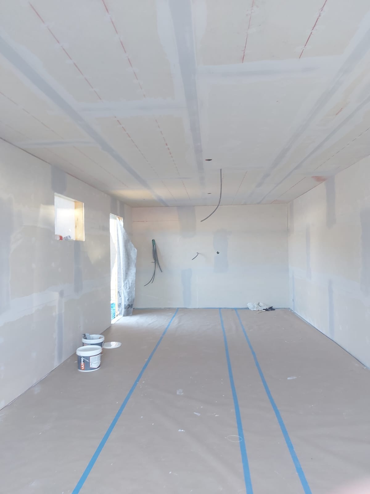 Wnętrze pokoju w trakcie prac wykończeniowych: ściany pokryte płytami gipsowo-kartonowymi ze śladami szpachlowania, podłoga zabezpieczona folią z niebieską taśmą, widoczne puszki z farbą.