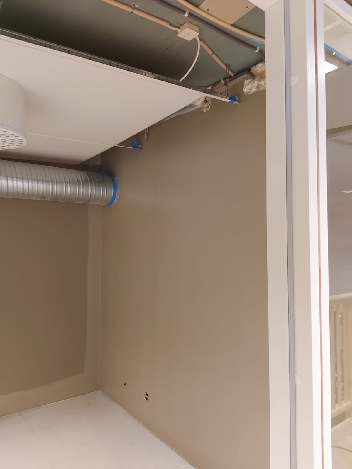 Świeżo pomalowane ściany w odcieniach beżu, widoczny fragment sufitu podwieszanego z wentylacją, narożnik pokoju i rama okienna.