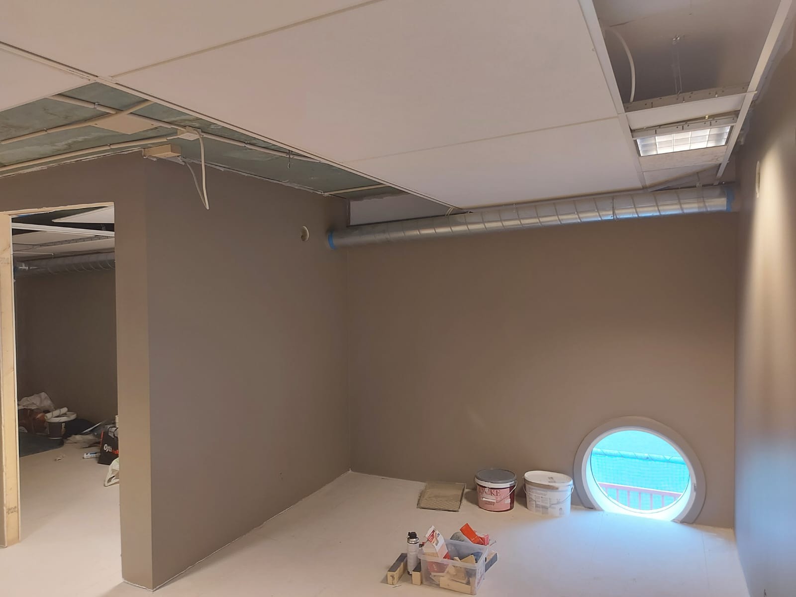 Wnętrze biura w trakcie prac wykończeniowych, ściany pomalowane na szaro, widoczne puszki z farbą, narzędzia malarskie, okrągłe okno z widokiem na zewnątrz, sufit podwieszany z otwartymi...