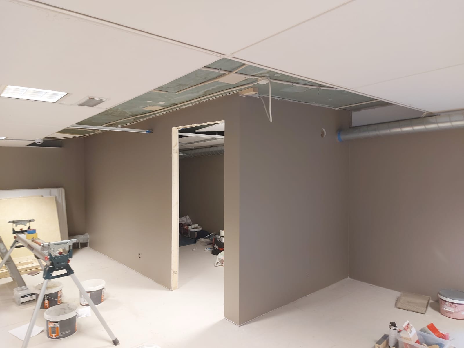 Przygotowane do malowania wnętrze biurowe z widocznymi śladami prac: drabina, puszki z farbą, szpachle i narzędzia malarskie. Ściany w kolorze beżowym, sufit w trakcie montażu.