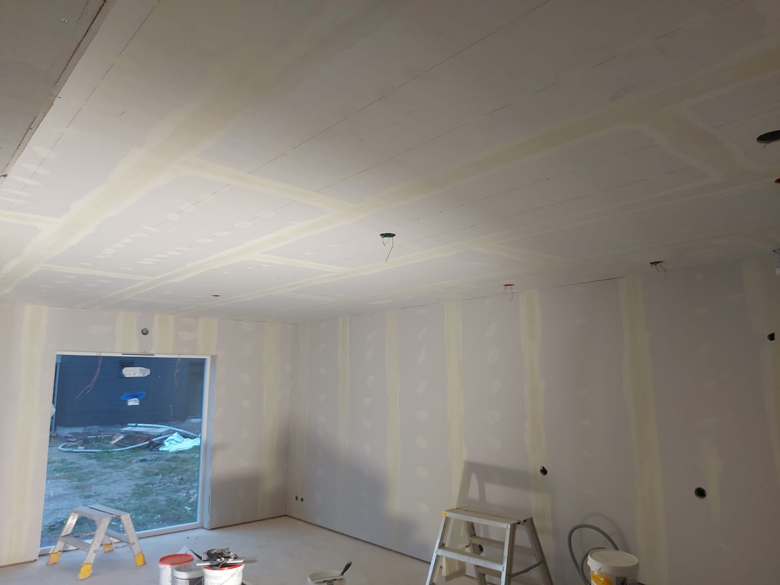 Wnętrze w Cerkwicy w trakcie prac wykończeniowych; widoczne ściany i sufit po szpachlowaniu, przygotowane do dalszych prac malarskich; na podłodze drabina i pojemniki z materiałami.