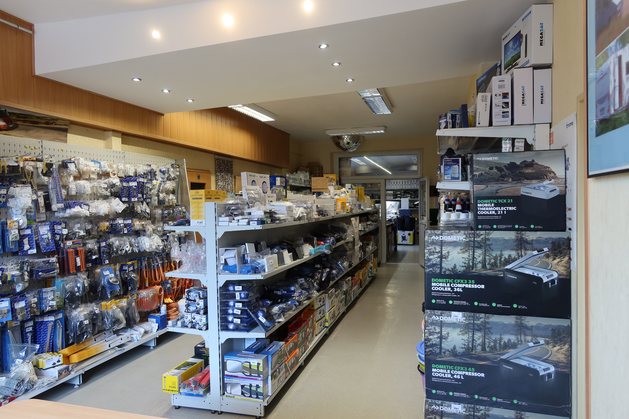 Wnętrze sklepu z akcesoriami i częściami do pojazdów kempingowych, widoczne regały z różnorodnym asortymentem, w tym chłodziarki kompresorowe Dometic w pudełkach.