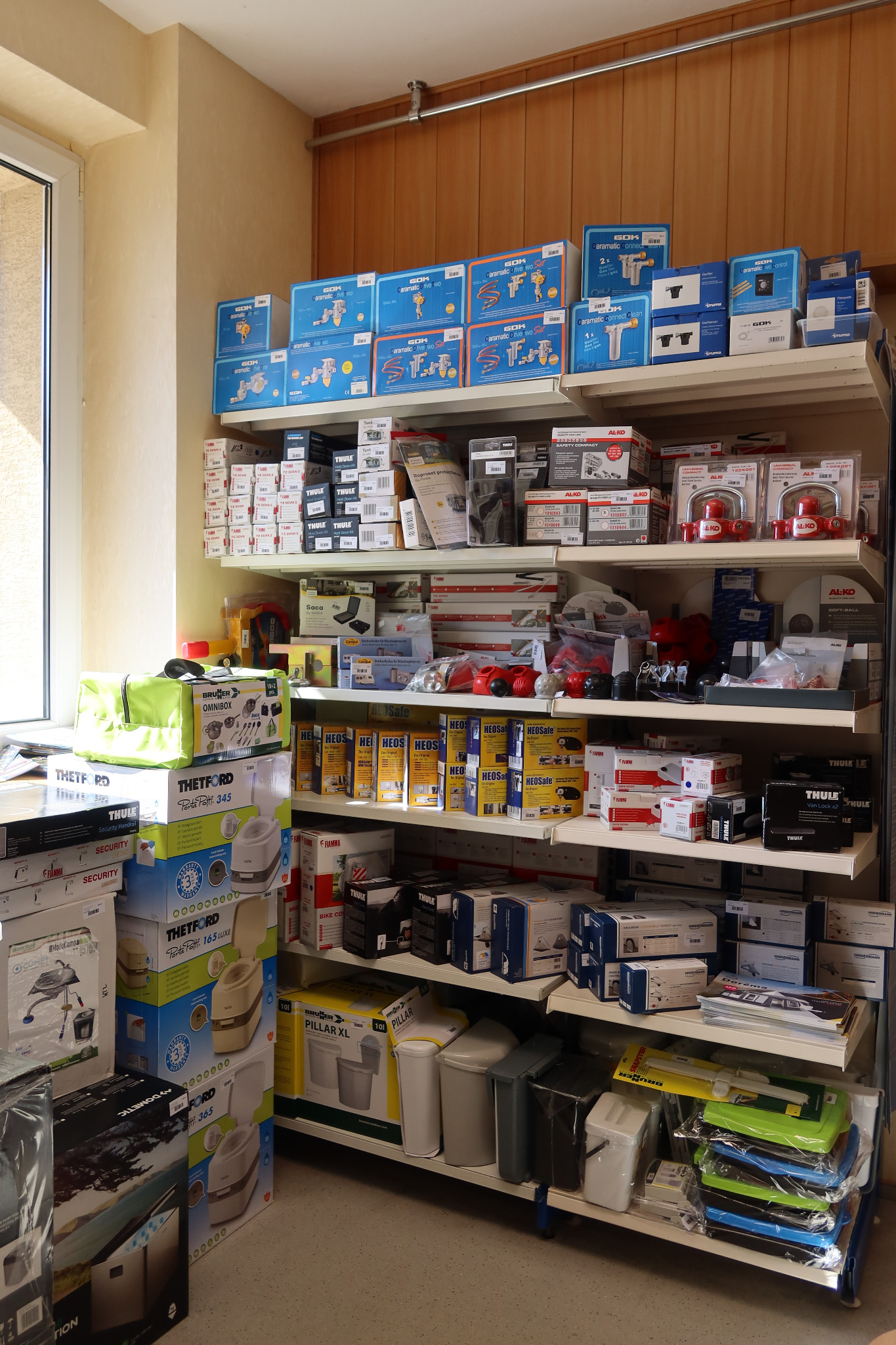 Półki sklepowe wypełnione różnorodnymi akcesoriami i częściami samochodowymi, głównie w oryginalnych opakowaniach, widoczne marki takie jak Thule, Thetford, Al-Ko i Heos, oświetlone naturalnym...