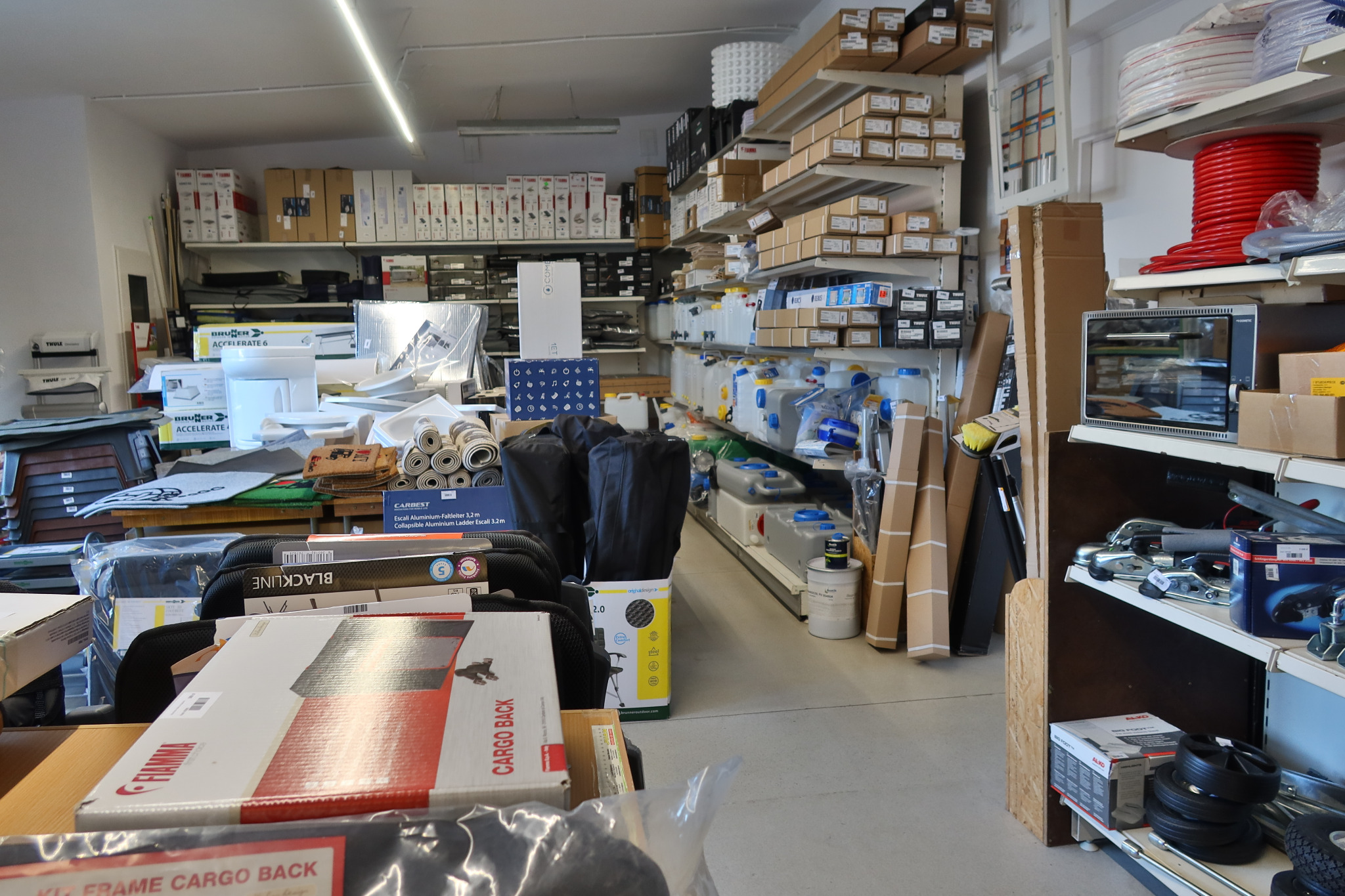 Wnętrze sklepu z akcesoriami samochodowymi i kempingowymi: regały wypełnione pudełkami, opakowaniami, zbiornikami na wodę i innymi produktami, widoczne marki Thule, Brunner, Fiamma i Carbest.