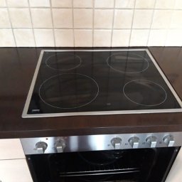 Monika Hinz - Kuchenka elektryczna z płytą indukcyjną, widok z góry na cztery pola grzejne, dolna część z otwartymi drzwiczkami piekarnika, tło stanowią jasne płytki ceramiczne.