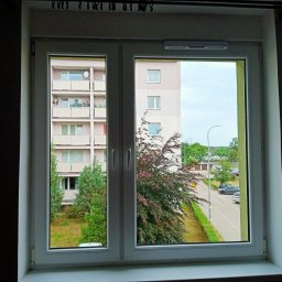 Monika Hinz - Widok z okna na blok mieszkalny z balkonami i ulicę z zaparkowanymi samochodami, widziany przez czyste, białe okno PCV.