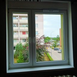 Monika Hinz - Widok z okna mieszkania na blok z balkonami i ulicę z samochodami w pochmurny dzień.