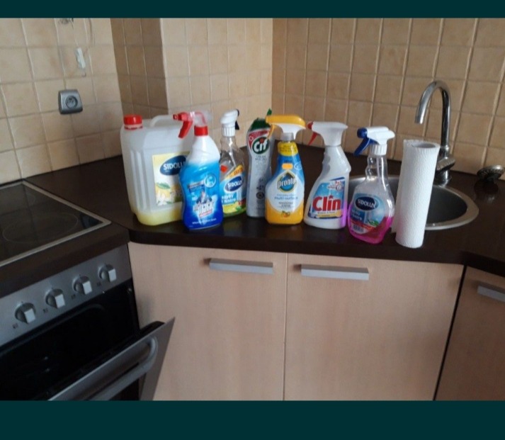 Różnorodne środki czystości, w tym Sidolux, Cif i Clin, ustawione na blacie kuchennym obok zlewu i piekarnika z otwartymi drzwiczkami.