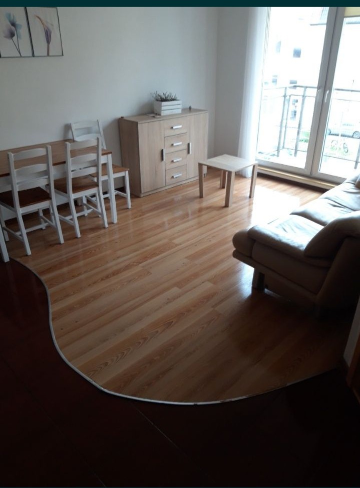 Jasne panele podłogowe w salonie z beżową sofą, drewnianym stołem i krzesłami oraz szafką z dekoracjami, widok na balkon.