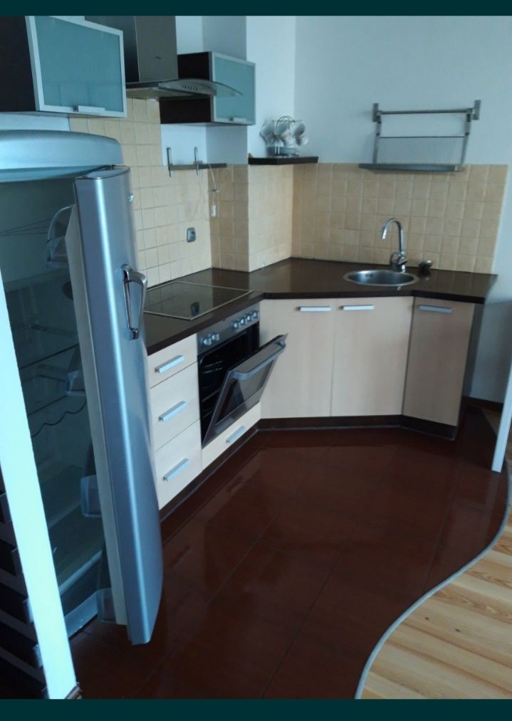 Kompaktowa kuchnia z beżowymi szafkami, ciemnym blatem i brązową podłogą, widoczny otwarty piekarnik i srebrna lodówka.