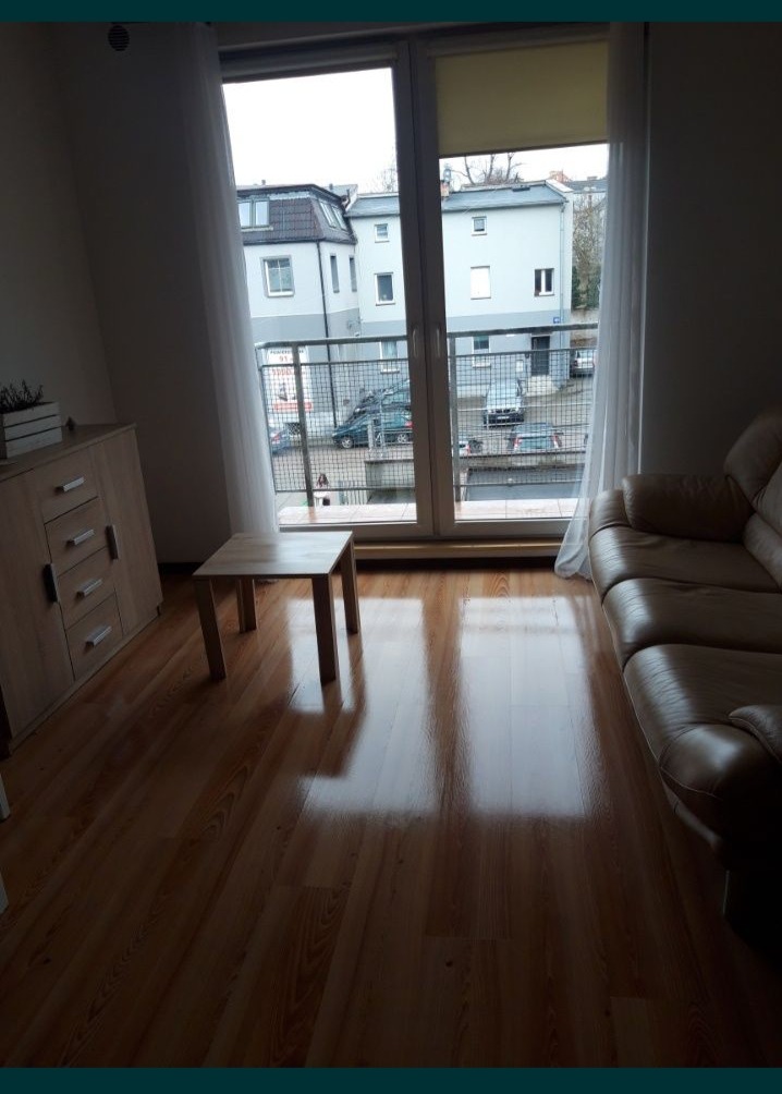 Jasne wnętrze pokoju z parkietem, beżową sofą i widokiem na balkon z budynkami w tle przez okno balkonowe.