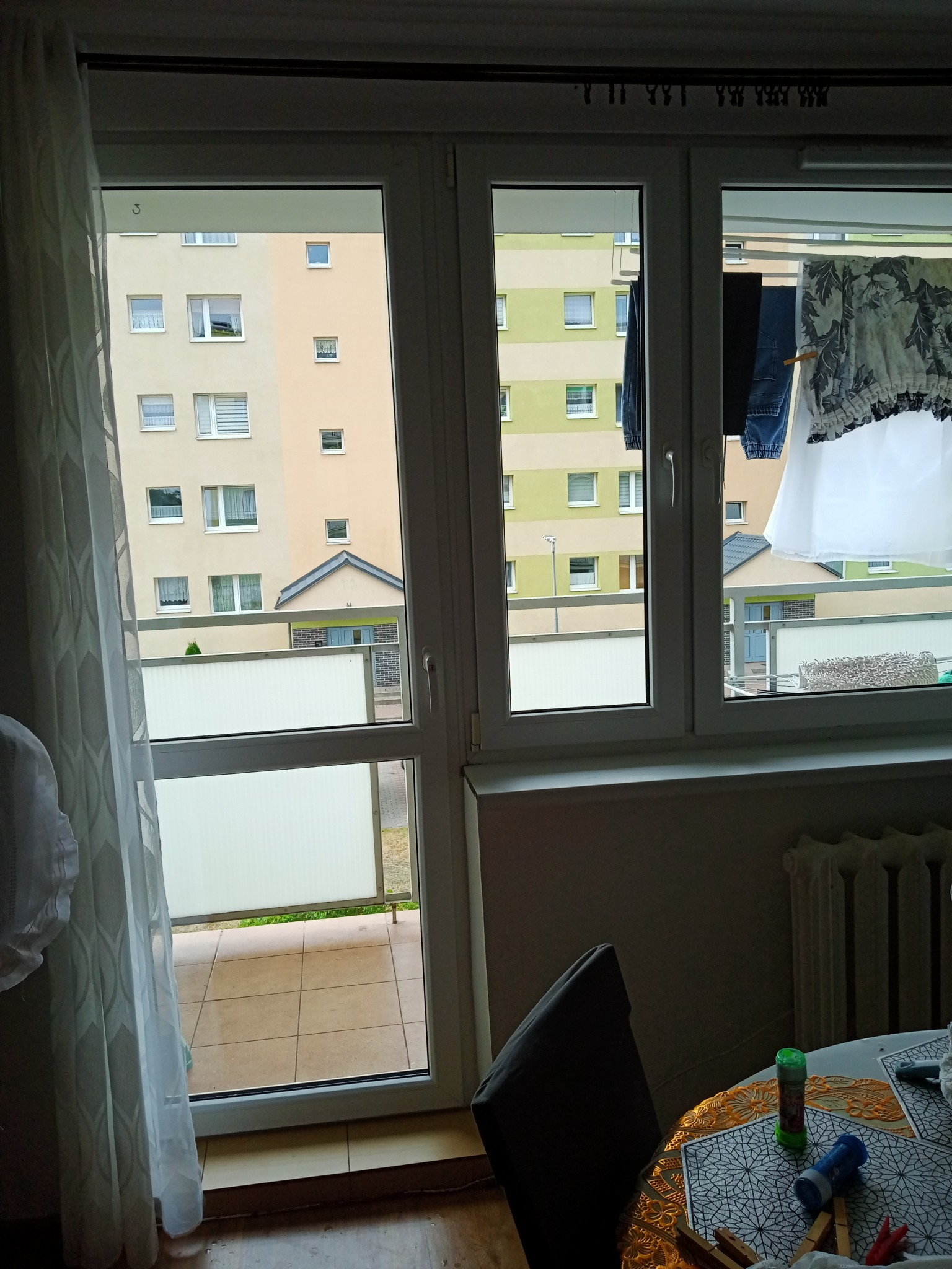 Widok z okna na suszące się pranie na balkonie w bloku, fragment stołu z płynem do baniek mydlanych w pomieszczeniu.