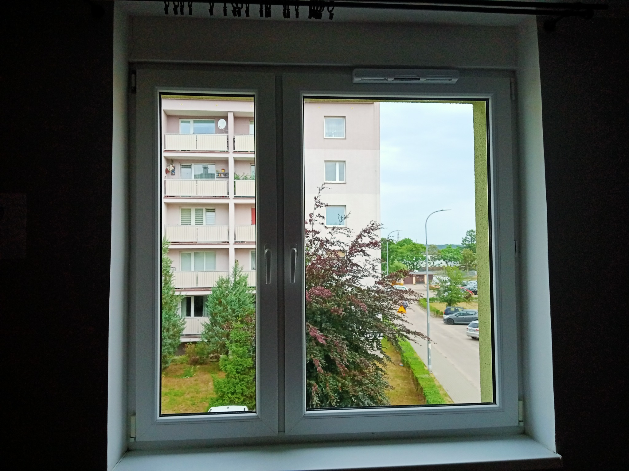 Widok z okna na blok mieszkalny z balkonami i ulicę z zaparkowanymi samochodami, widziany przez czyste, białe okno PCV.